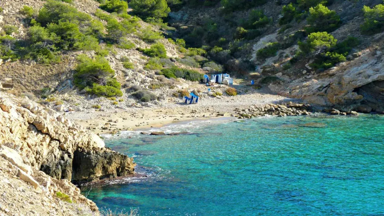 Agua turquesa en la cala Almadrava de Benidorm, con rocas y vegetación en la ladera. Hay algunas sillas y sombrillas en la playa.