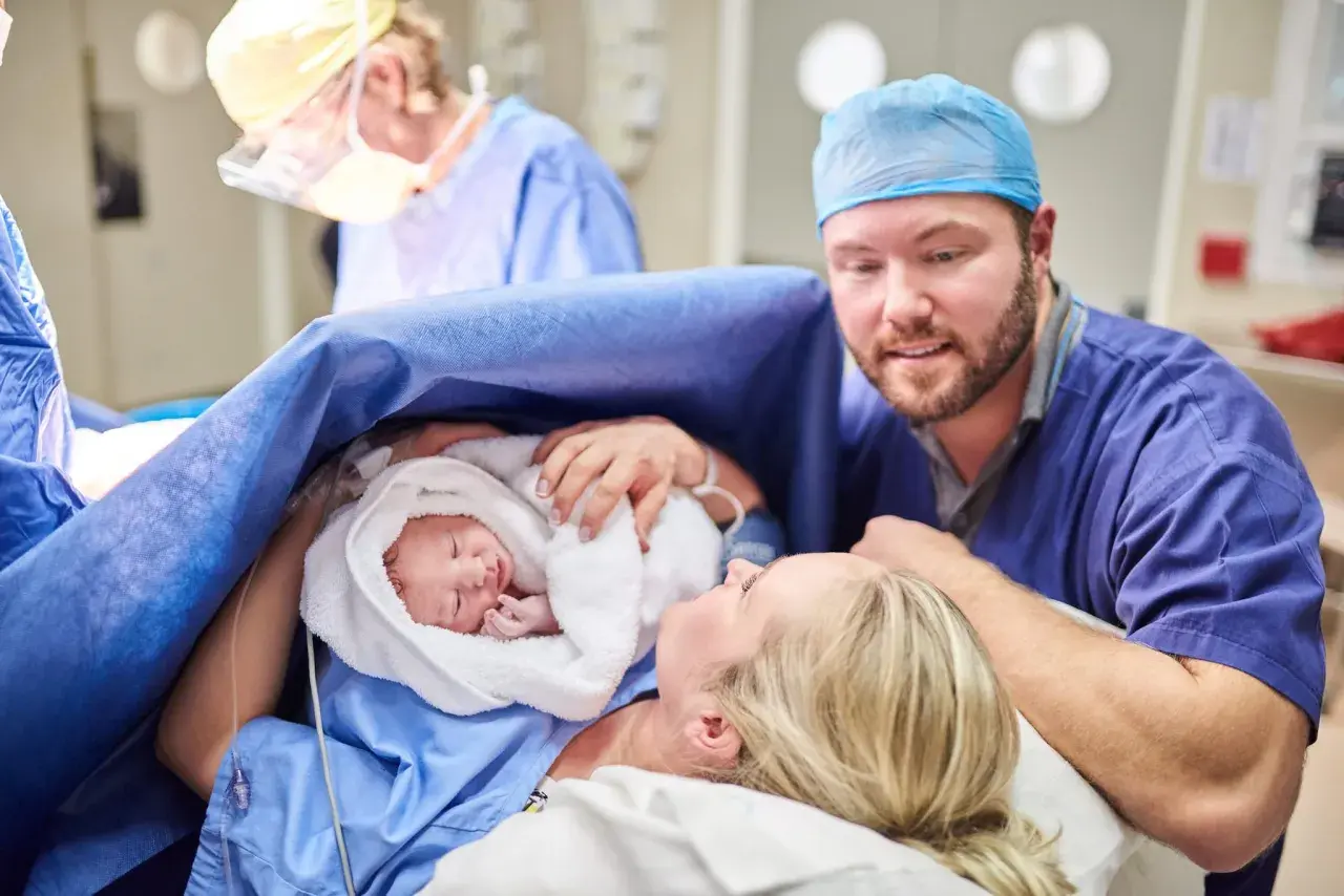 Un padre guarda con amore il suo neonato, avvolto in un asciugamano bianco, dopo un cesareo dolce. La madre sorride, circondata da personale medico.