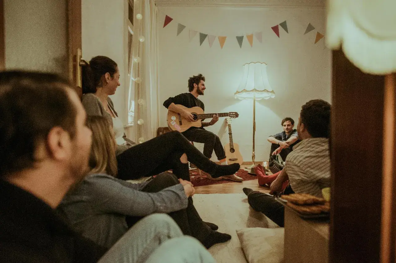 Freunde versammeln sich für einen gemütlichen Abend mit Gitarrenmusik und **Lieder zum Mitsingen**. Bunte Wimpelketten schmücken die Wand.