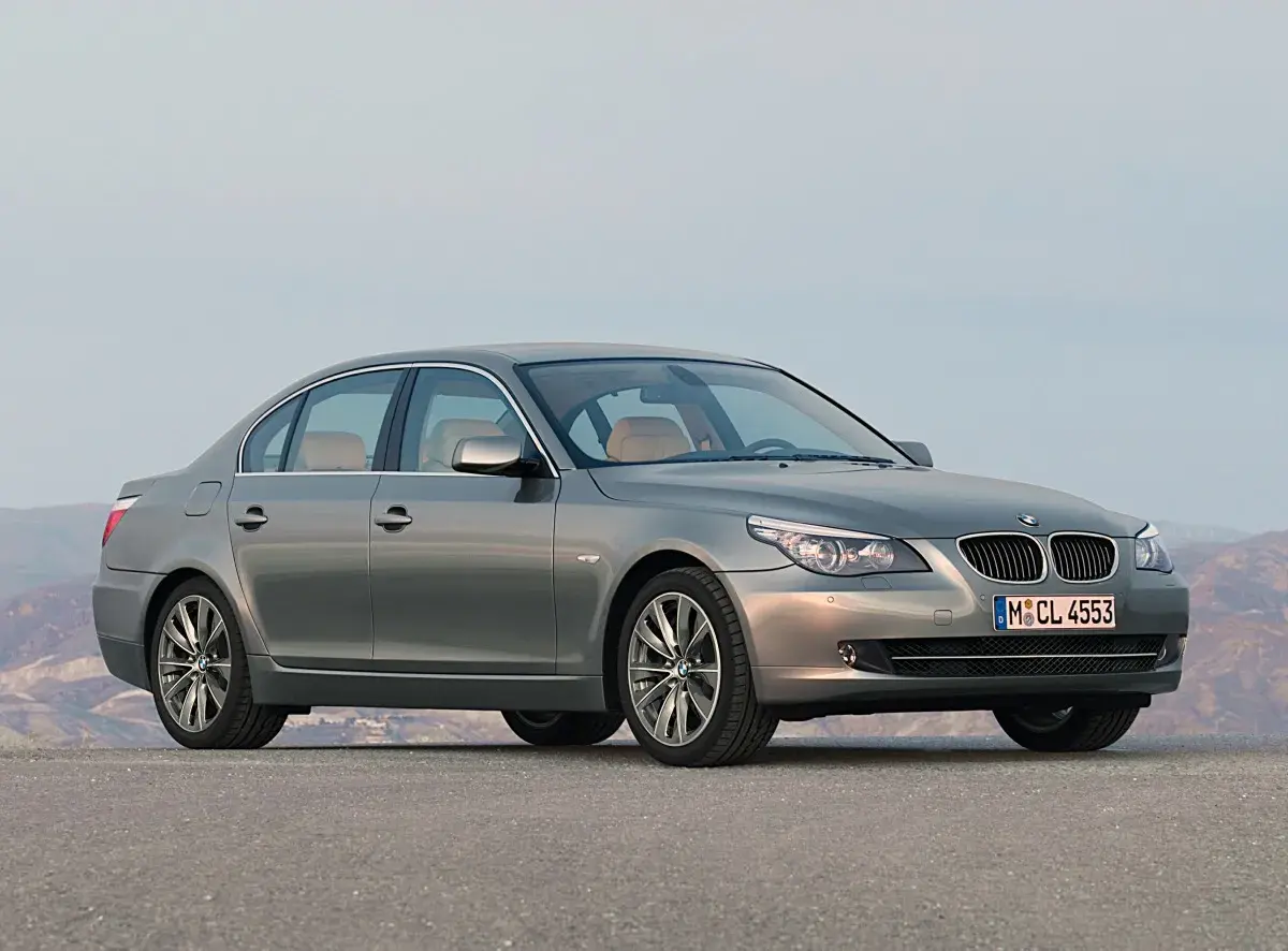Szary sedan BMW 5 Series (E60) z jasnym wnętrzem stoi na pustej drodze, z górami w tle.