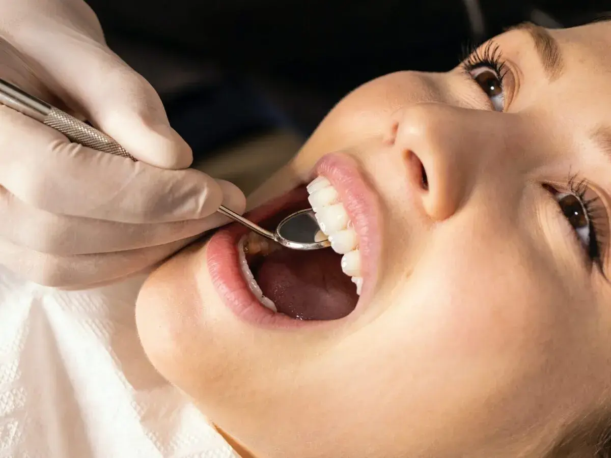 Uśmiechnięta pacjentka podczas zabiegu plastyka dziąseł. Dentysta bada zęby lusterkiem. Efekt plastyka dziąseł przed i po jest widoczny.