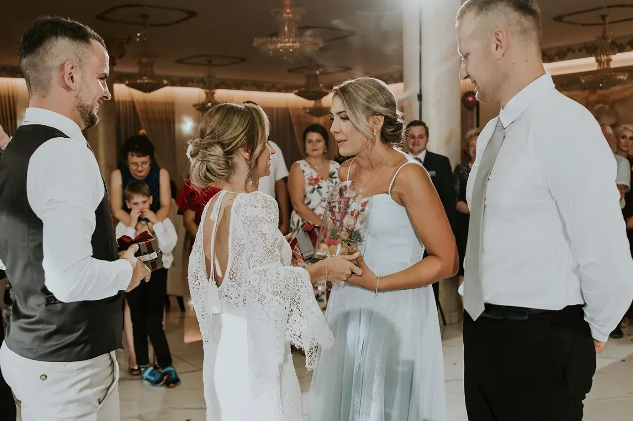 Nietypowe podziękowania dla rodziców na weselu: panna młoda w koronkowej sukni wręcza bukiet w szkle, a pan młody prezent z wstążką.