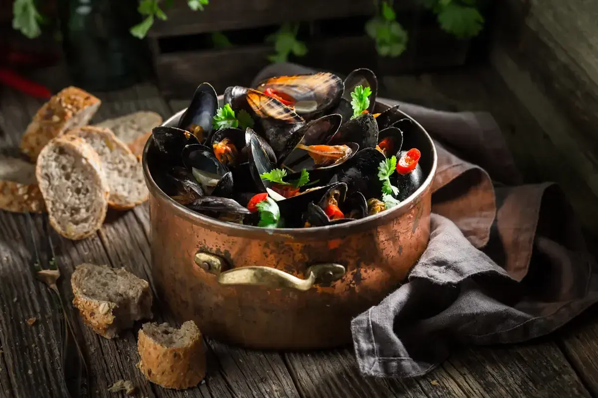 Un grand pot en cuivre rempli de moules ouvertes avant cuisson, garnies de persil et de piment. Du pain rustique est disposé sur la table en bois.