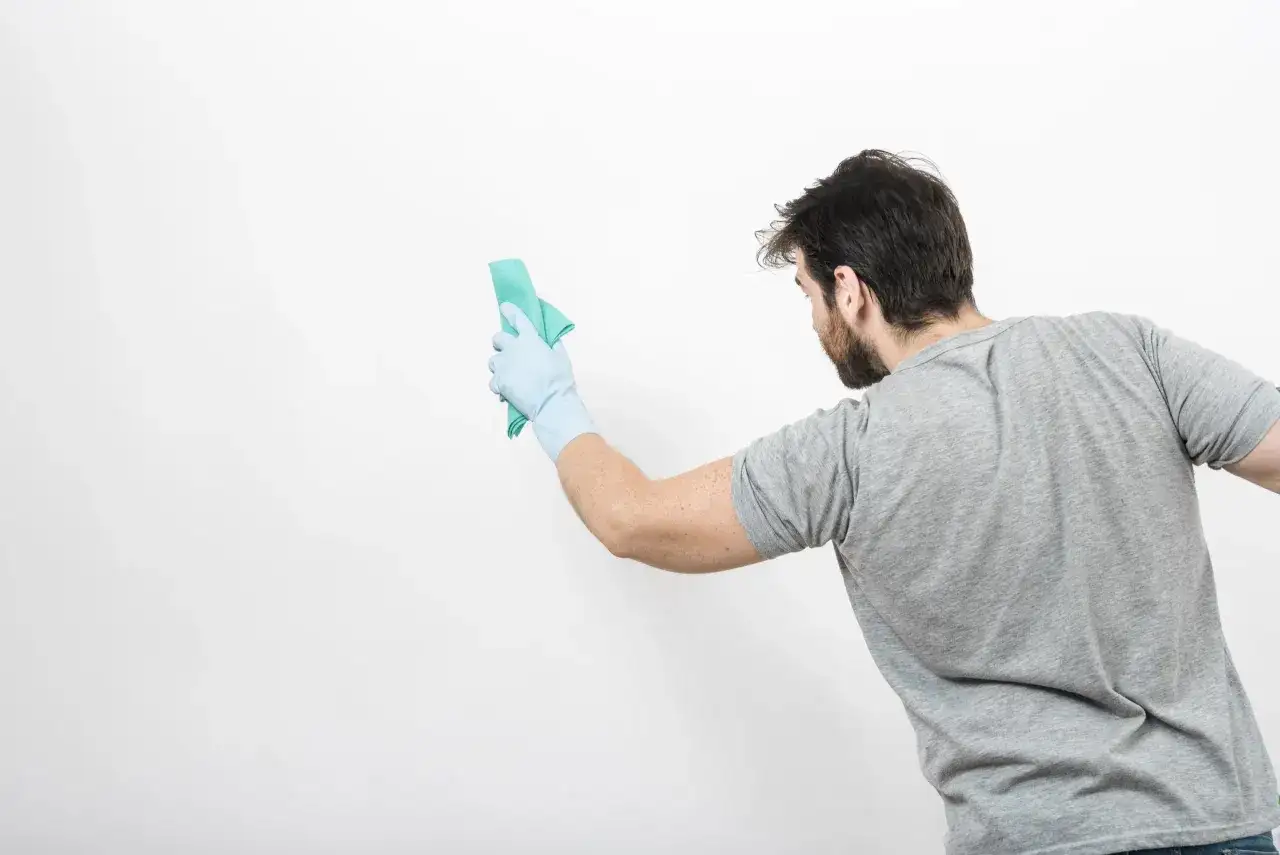 Un homme en gant bleu nettoie un mur blanc avec un chiffon vert. Il montre comment nettoyer un mur blanc sans enlever la peinture.