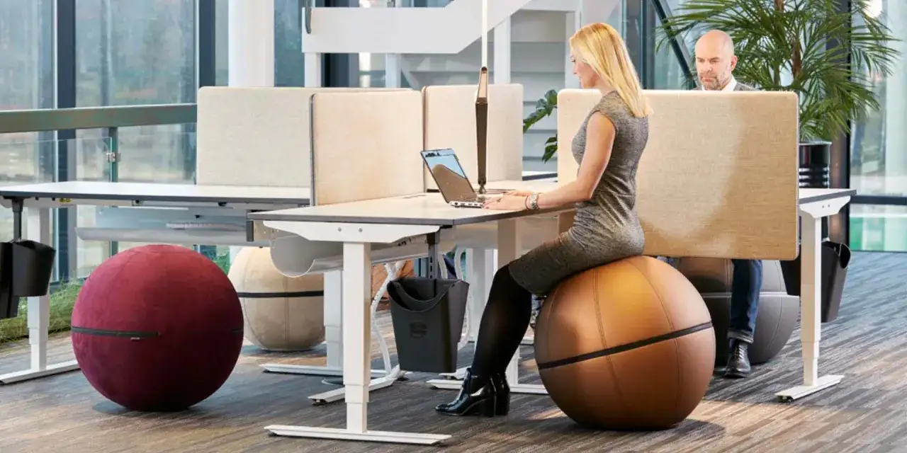 Kvinna arbetar vid ett höj- och sänkbart skrivbord i ett aktivitetsbaserat kontor. Hon sitter på en stor, brun träningsboll.