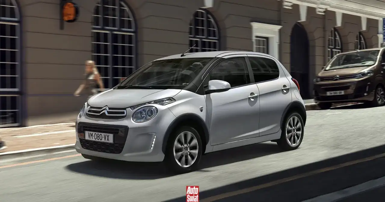 Srebrny Citroën C1 z czarnymi nakładkami na błotniki przemierza miejską ulicę.
