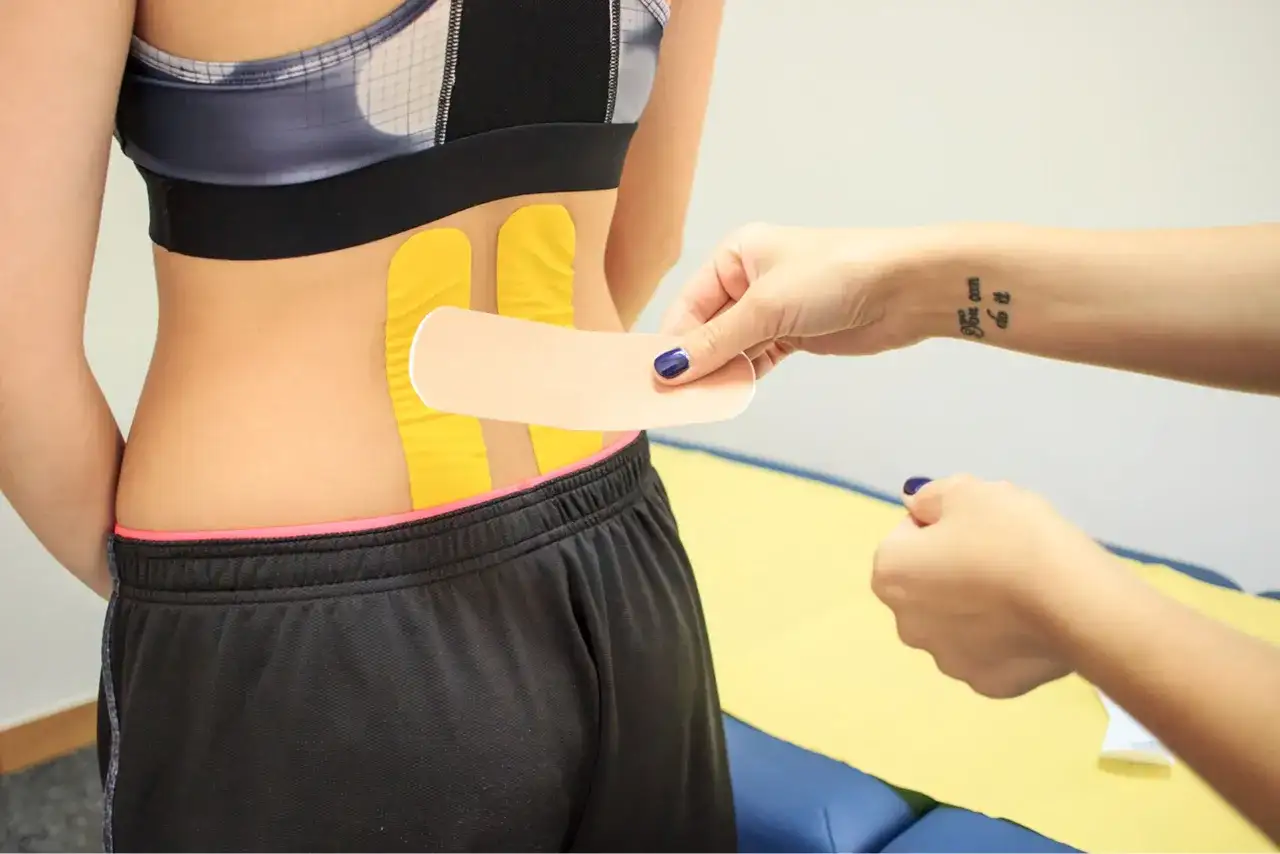 Naklejanie tejpy na plecy w odcinku lędźwiowym. Dłoń z tatuażem "You can do it" przygotowuje plaster.