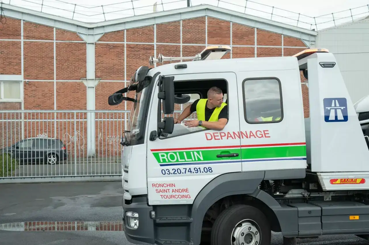 Un dépanneur en uniforme jaune regarde par la fenêtre d'un camion de dépannage. Le camion est marqué "ROLLIN DEPANNNAGE".