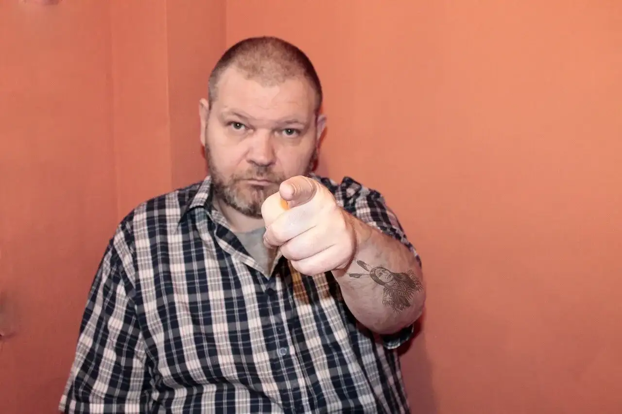 Mężczyzna w kratę wskazuje palcem, na ramieniu tatuaż z Indianinem. "234 kk" - czy to jego numer?