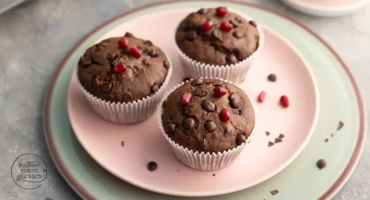 Drei saftige, vegane Muffins mit Schokostückchen und Granatapfelkernen auf einem rosa Teller.