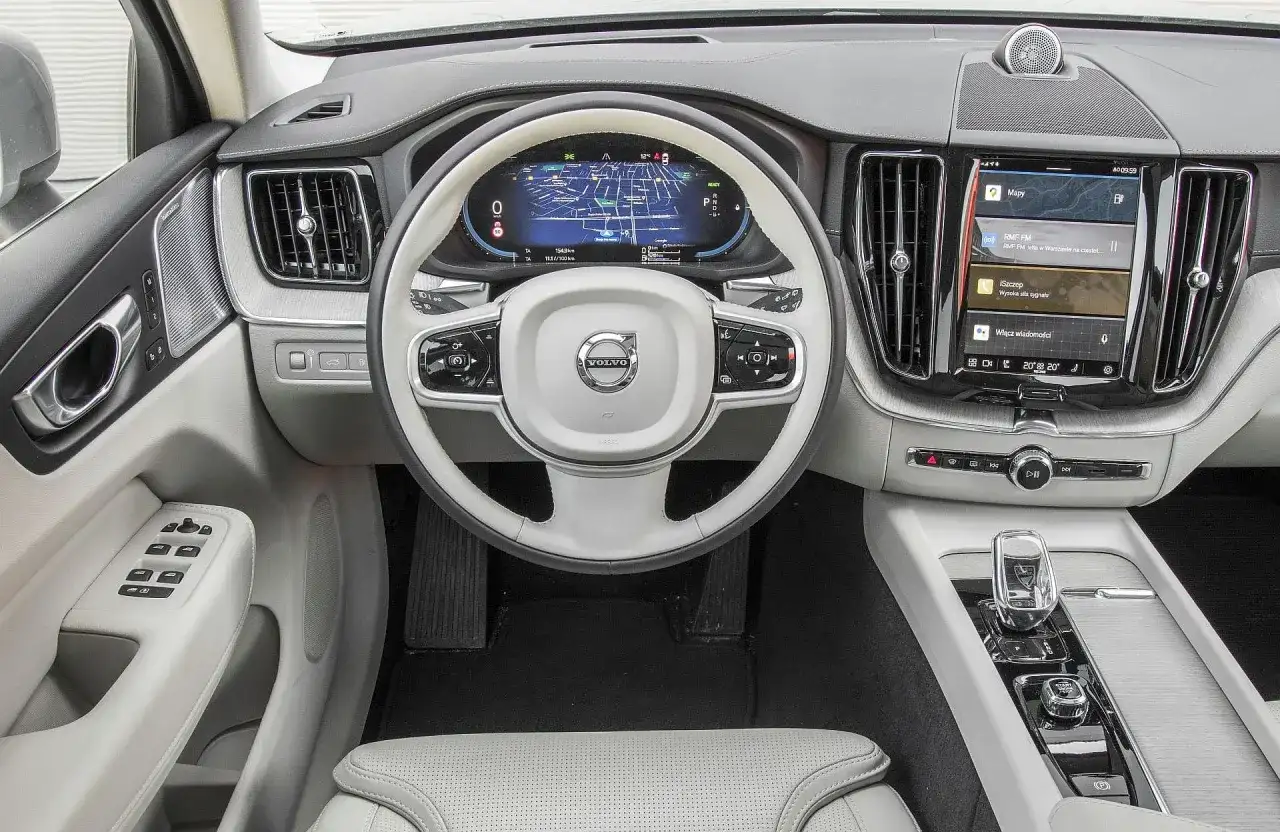 Wnętrze Volvo XC60 T6 z ekranem nawigacji i multimediów. Jasna tapicerka, biała kierownica z logo Volvo.