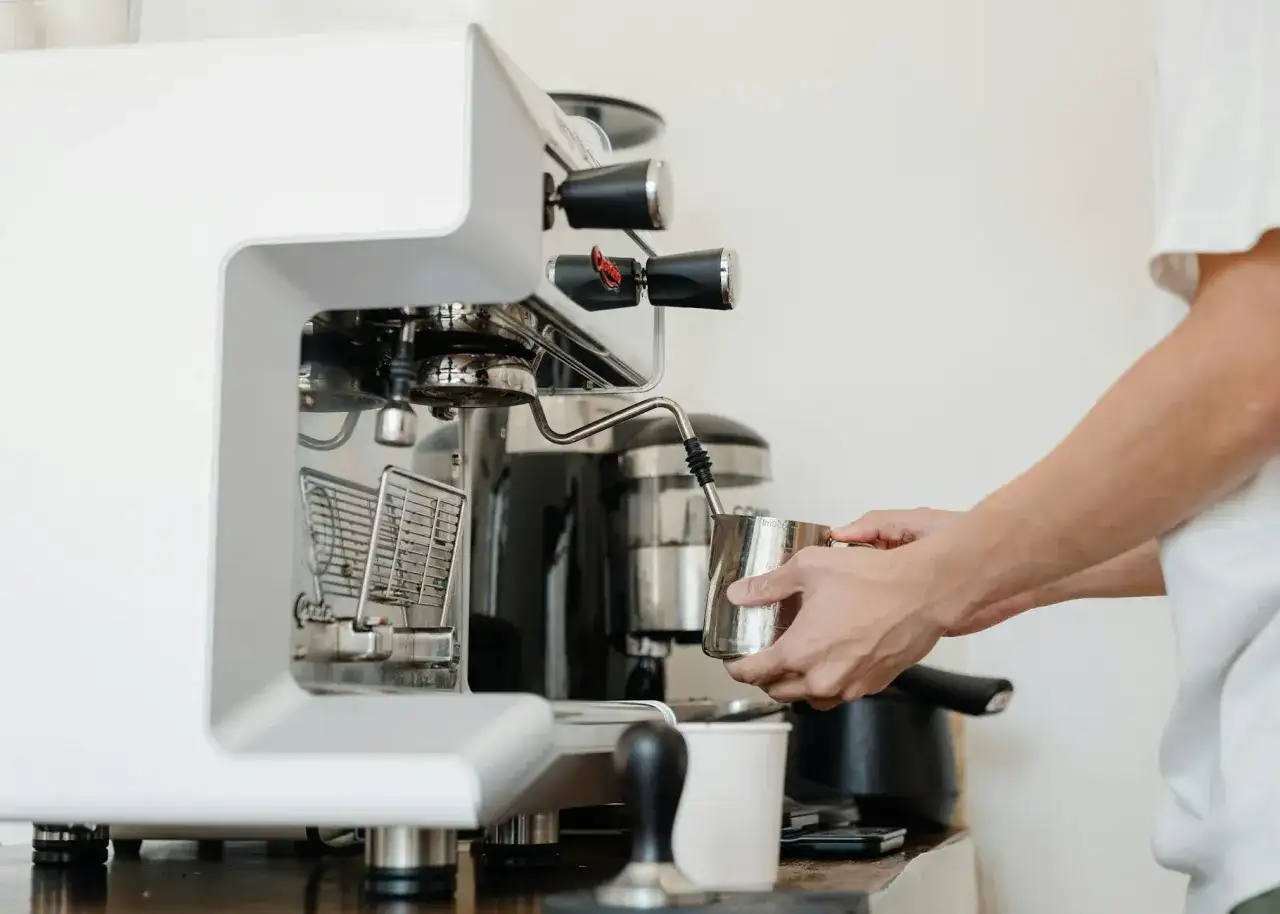Barista spienia mleko w dzbanku, używając dyszy ekspresu. W tle widoczny blok zaparzający.