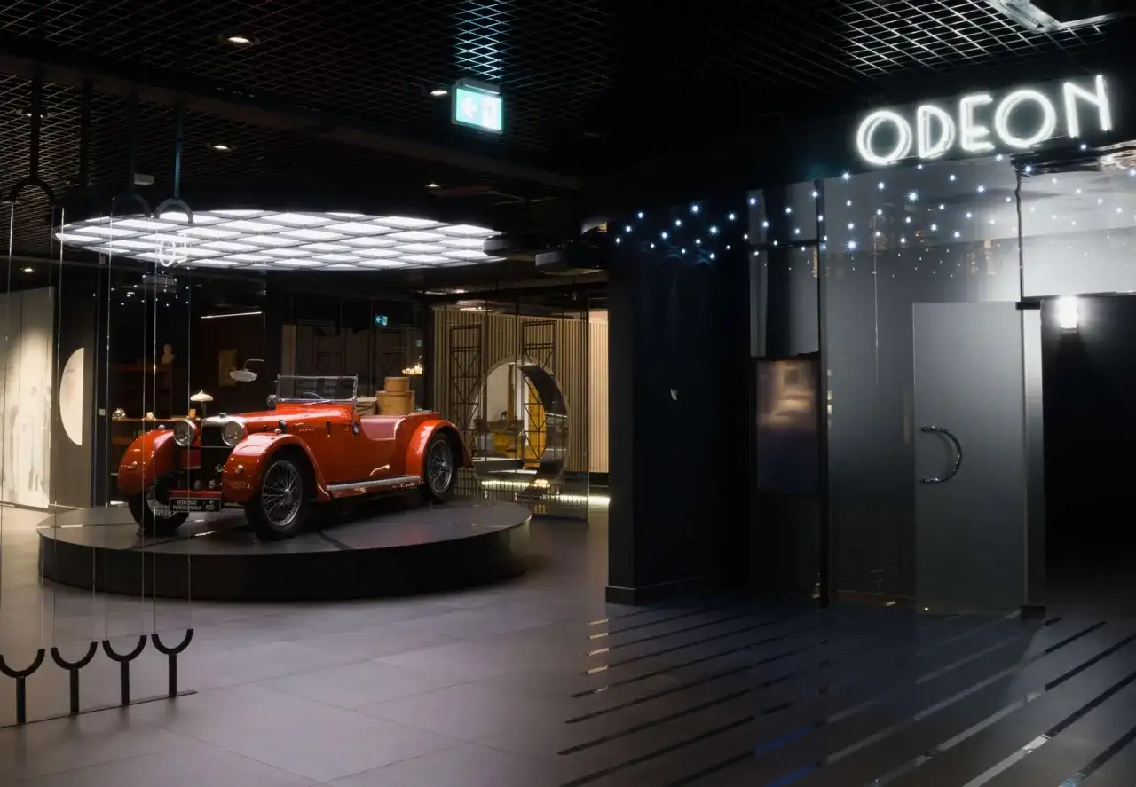 Pomarańczowy kabriolet z lat 20. XX wieku, symbol sztuki dwudziestolecia międzywojennego, prezentowany w nowoczesnym wnętrzu z neonowym napisem "ODEON".