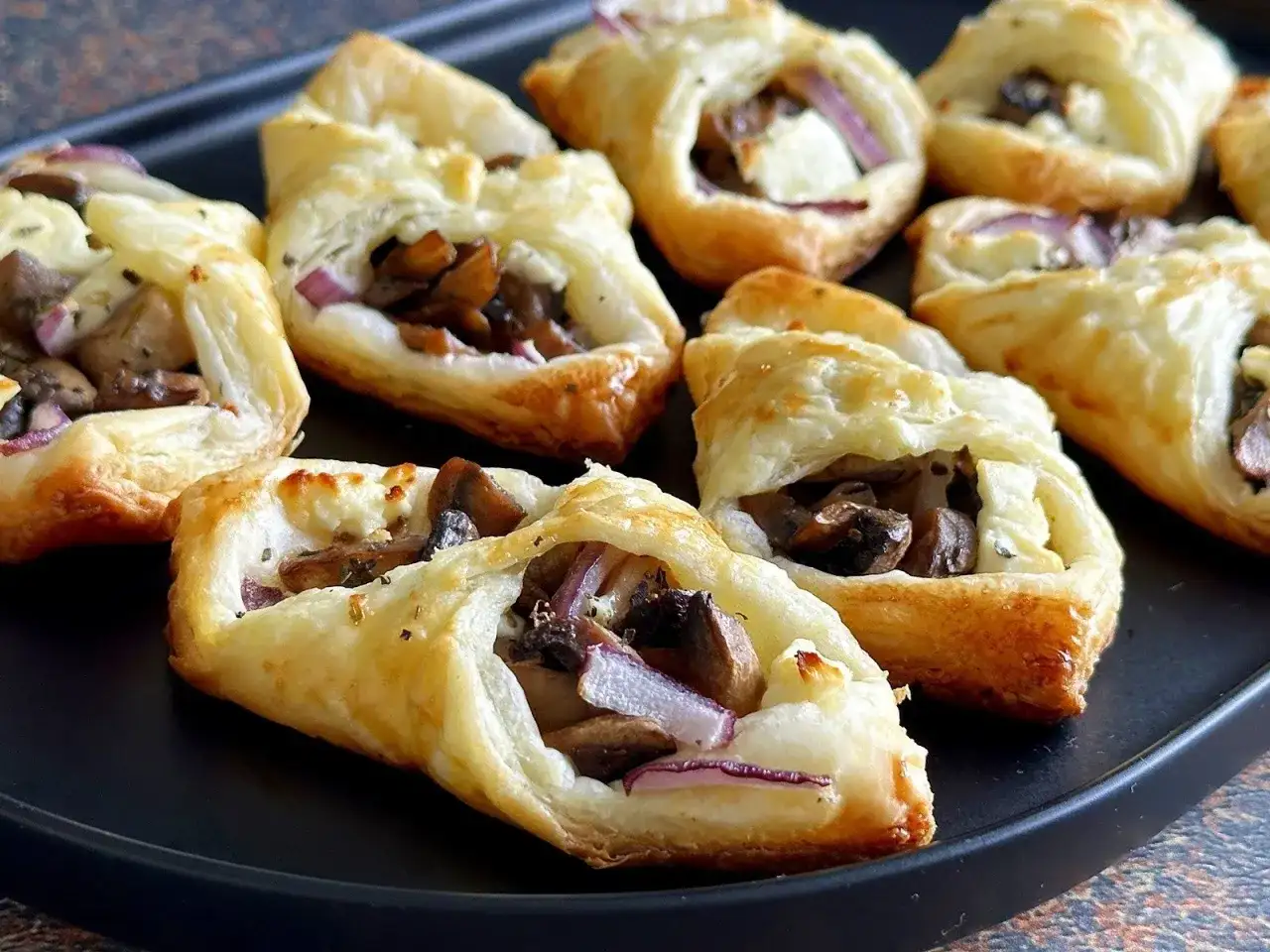 Chrupiące ciasto francuskie z pieczarkami, serem feta i czerwoną cebulą, ułożone na czarnej tacy.