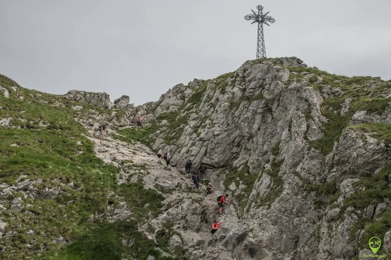 Grupa turystów wspina się po kamienistym szlaku na Giewont. W oddali widać charakterystyczny krzyż.