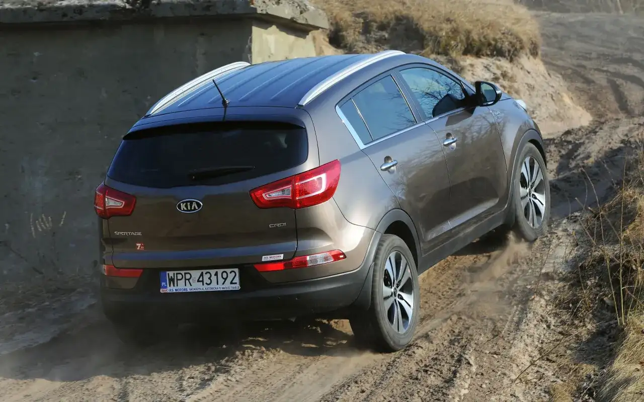 Brązowy Kia Sportage 2014 zjeżdża po piaszczystej ścieżce, unosząc kurz.