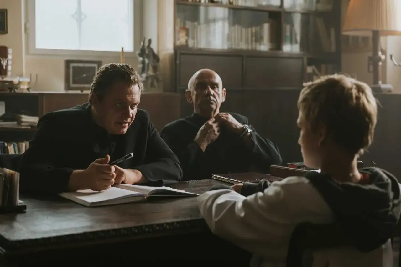 Trzech mężczyzn przy stole, jeden z nich pisze. Scena z filmów Piotra Domalewskiego.