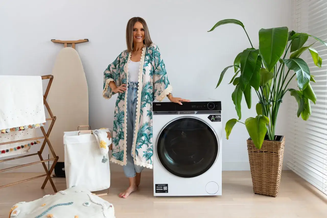 Kobieta w kwiecistej narzutce obok nowoczesnej pralki Haier. Wiele osób szuka pralki Haier opinie, by wybrać najlepszy model.