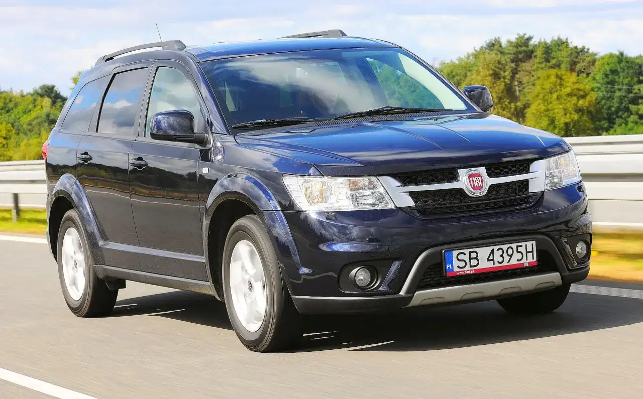 Ciemnoniebieski Fiat Freemont jedzie po drodze, otoczony zielenią i błękitnym niebem.
