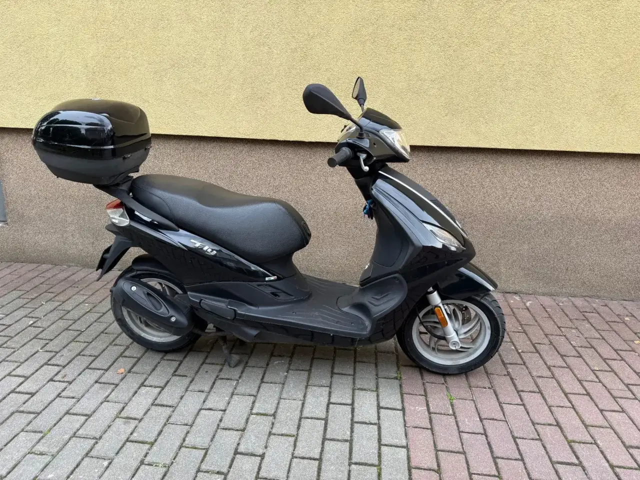 Czarny skuter Piaggio Fly 50 z kuferkiem na bagaż, zaparkowany na chodniku z kostki brukowej.