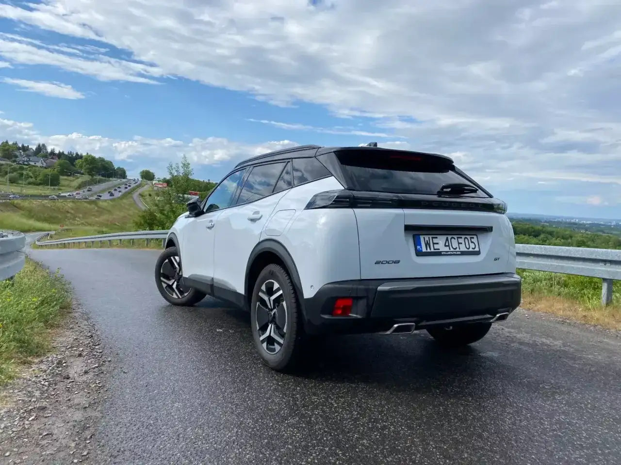 Biały Peugeot 2008 GT na mokrej drodze, z widokiem na krętą trasę i zielone wzgórza.