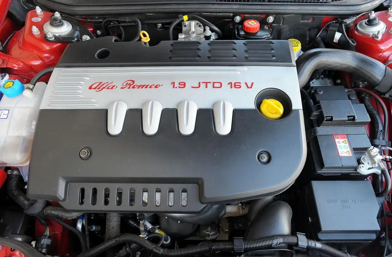 Silnik Alfa Romeo 147 1.9 JTD 16V, widok z góry. Czerwona karoseria, czarna osłona silnika z napisem.