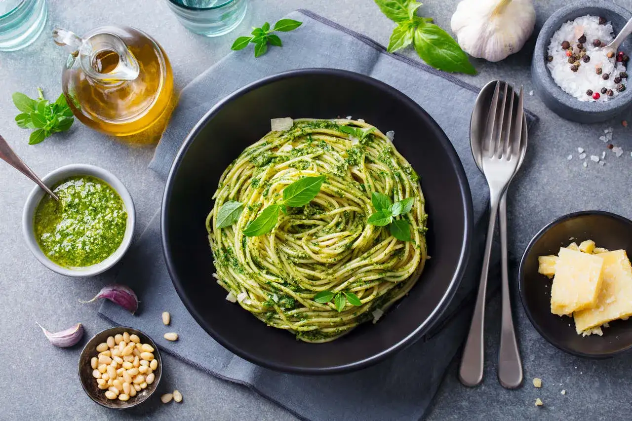 Spaghetti z pesto i bazylią w czarnej misce, obok sos, orzeszki piniowe, czosnek i parmezan.