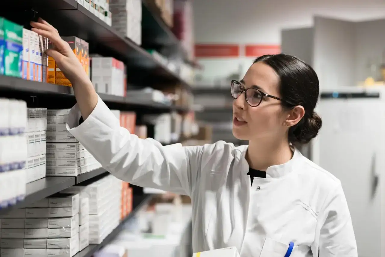 Farmaceutka w białym kitlu sięga po lek, być może pentoksyfilinę, z aptecznej półki pełnej opakowań.