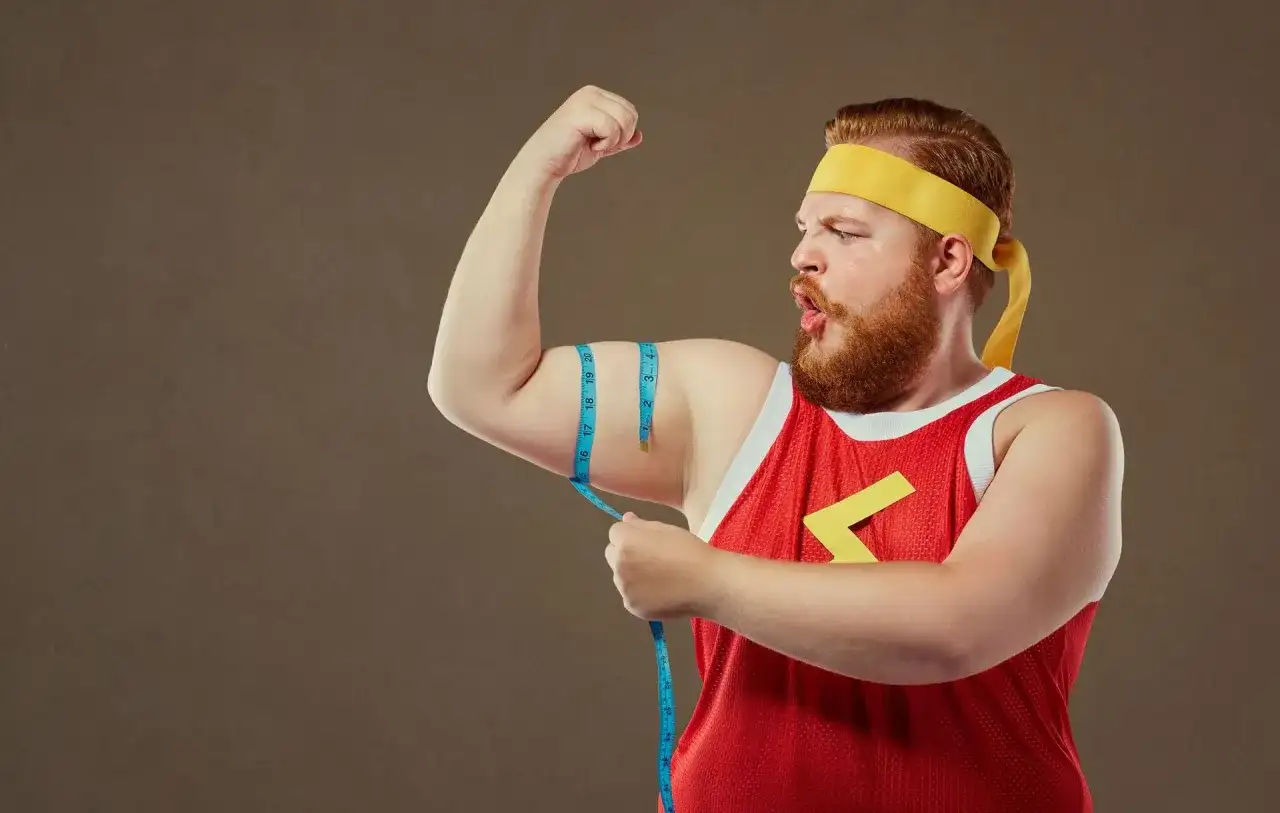 Rudowłosy mężczyzna typu endomorfik w żółtej opasce i czerwonej koszulce mierzy obwód bicepsa.