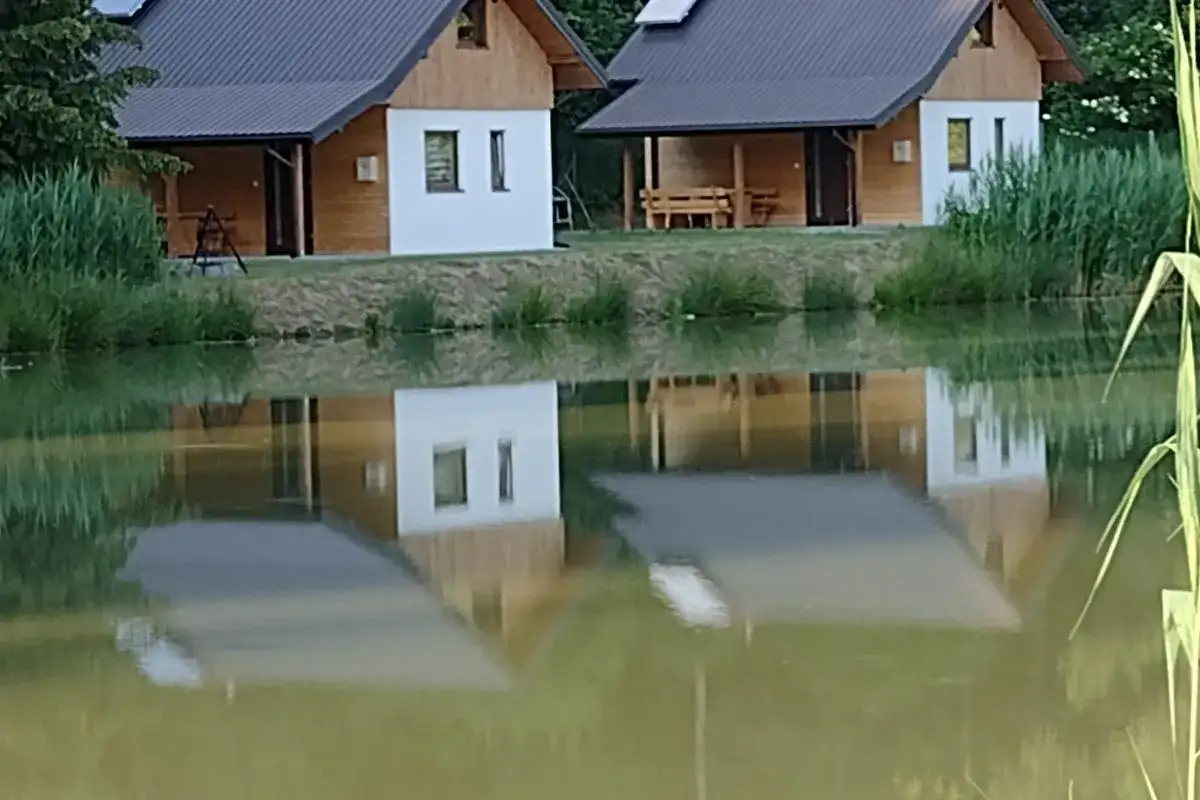 Urokliwe łowisko z domkami w Podkarpackiem. Dwa drewniano-białe domki z dachami odbijają się w spokojnej tafli wody.