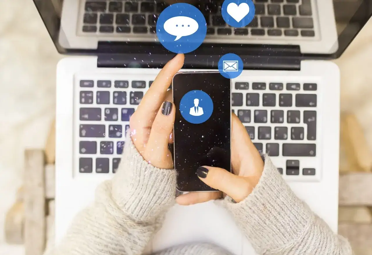 Jak zobaczyć czy ktoś cię zablokował na Messengerze? Dłonie trzymające smartfon nad klawiaturą laptopa, ikony czatu, serca i wiadomości.