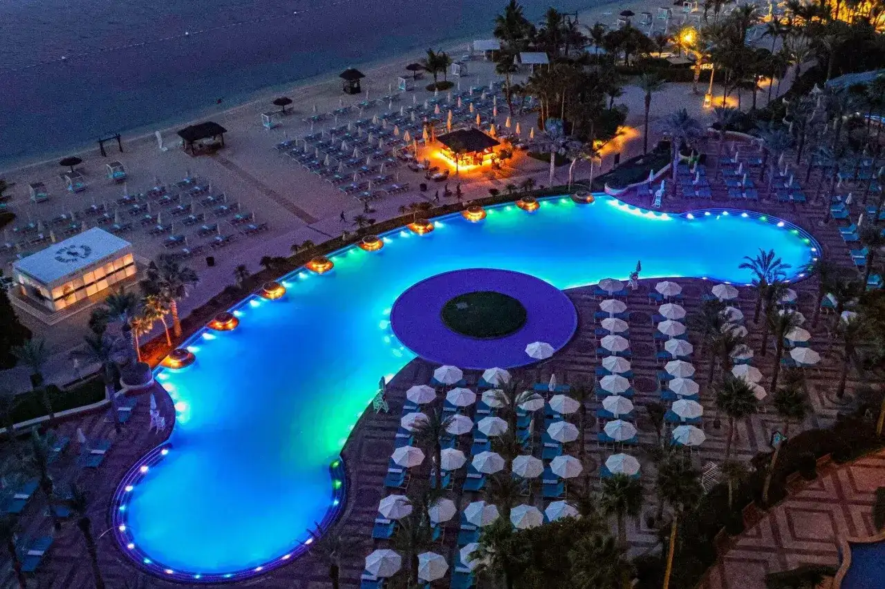 Nocny widok na luksusowy basen i plażę w Dubaju. Błękitna woda basenu, oświetlona kolorowymi światłami, otoczona leżakami i palmami.
