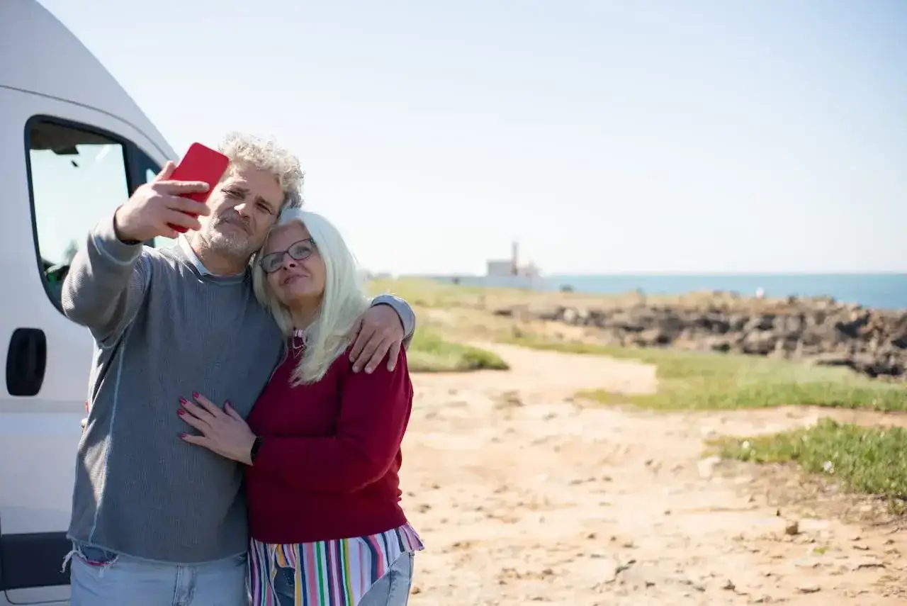 Para singli po 50-tce, którzy szukają przygód, idealne są wycieczki kamperem. Para robi sobie selfie na tle morza.