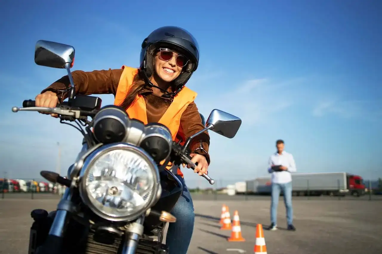 Uśmiechnięta kursantka w kamizelce i kasku na motocyklu, gotowa do zdobycia prawa jazdy na motocykl. Instruktor obserwuje w tle.