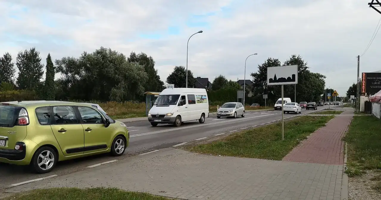 Zielony samochód i biały bus na drodze w terenie zabudowanym. W tle drzewa i domy.