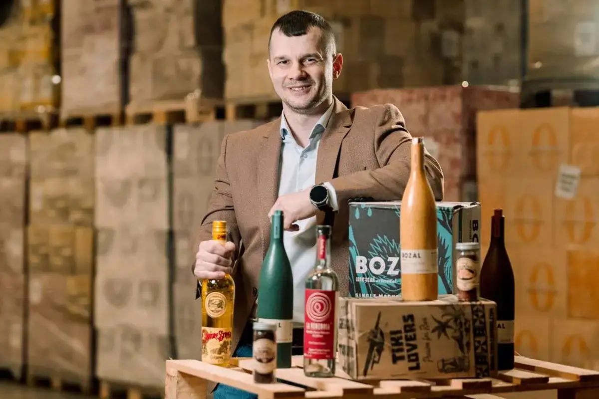 Mężczyzna w brązowej marynarce prezentuje butelki z alkoholem, w tym te z etykietą "Spice Bros". W tle magazyn pełen towarów, sugerujący dystrybucję california alkohol.