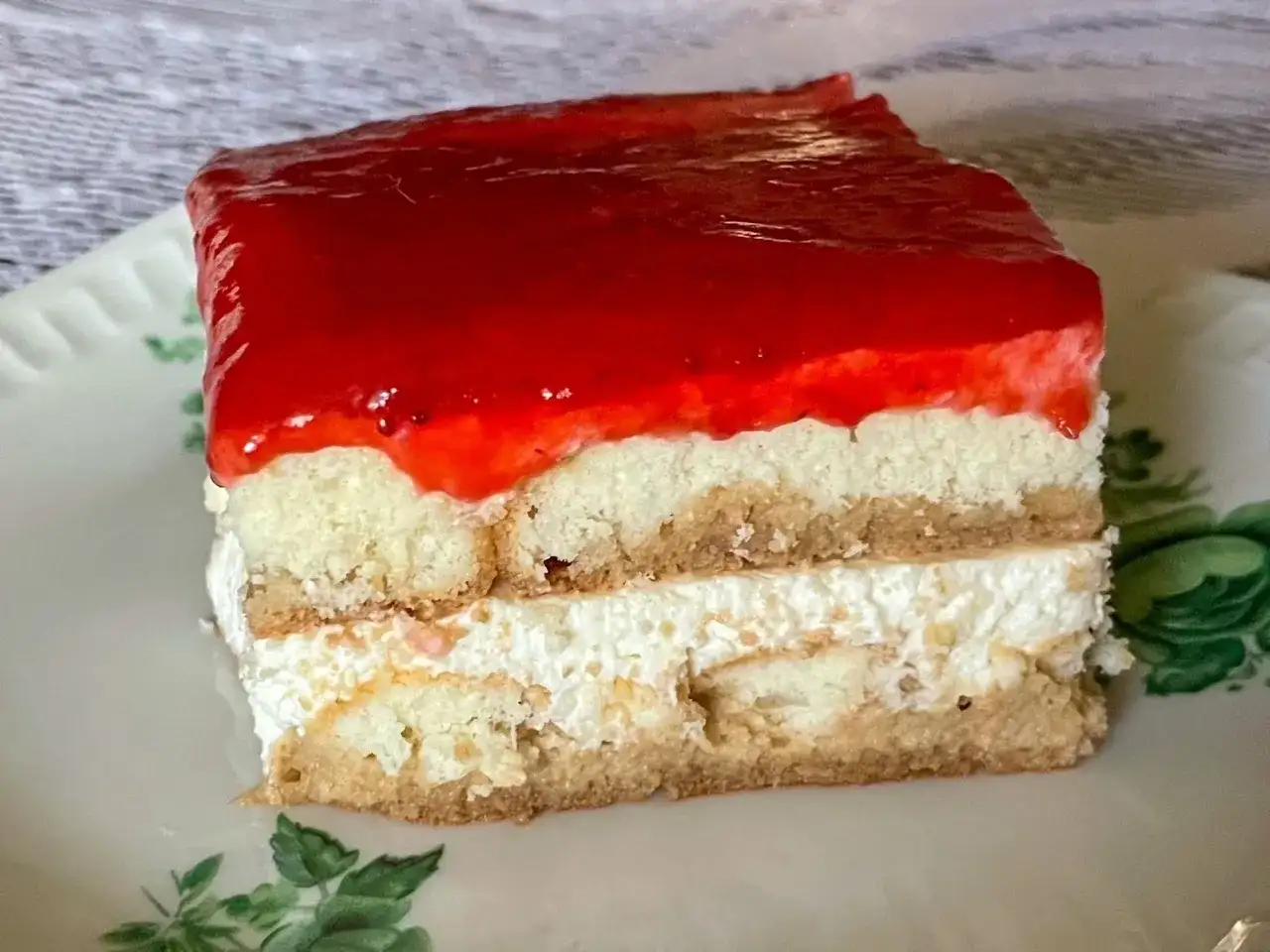 Kawałek ciasta bez pieczenia z warstwami biszkoptu, kremu i galaretki truskawkowej na talerzyku.
