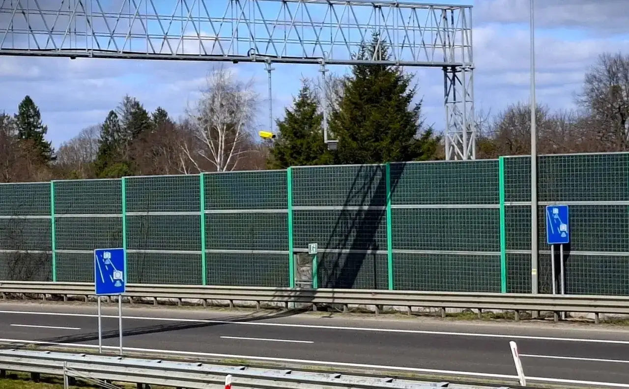 Znaki informują o odcinkowym pomiarze prędkości. Za zieloną barierą dźwiękochłonną widać drzewa i niebo.
