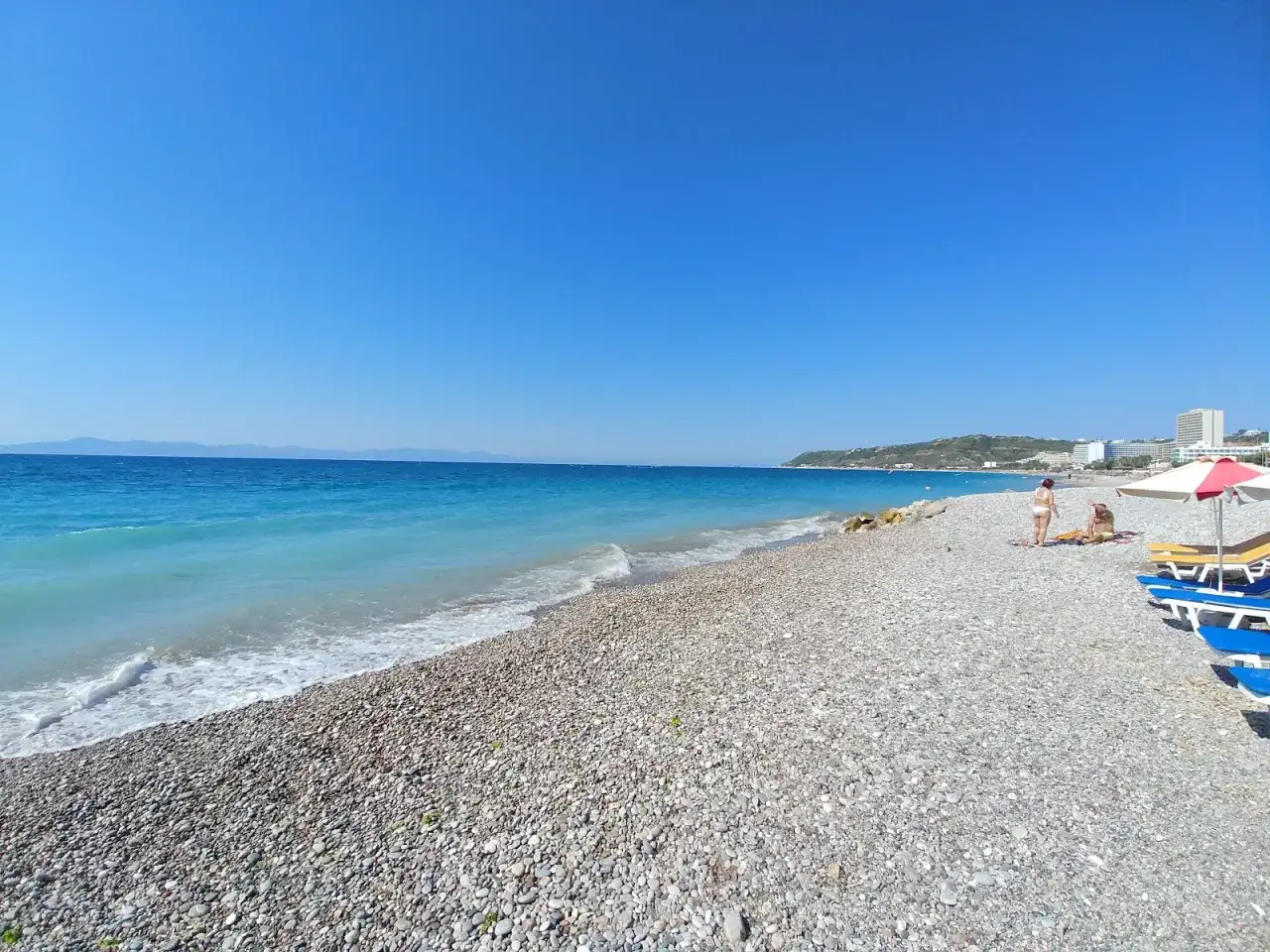 Słoneczna plaża w Ixia, Rodos, z turkusowym morzem i kamienistym brzegiem. Dwie osoby odpoczywają pod parasolem.