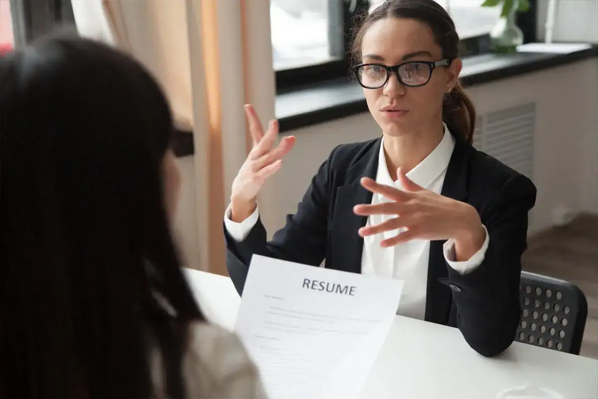 Kobieta w okularach i czarnej marynarce prowadzi rozmowę kwalifikacyjną, gestykulując rękami nad dokumentem "RESUME".