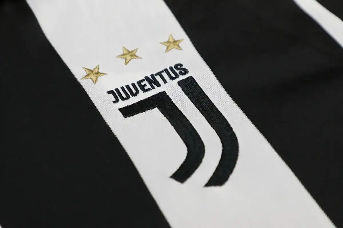 Zbliżenie na herb Juventusu z trzema złotymi gwiazdkami i napisem "JUVENTUS" na tle charakterystycznych czarno-białych pasów.