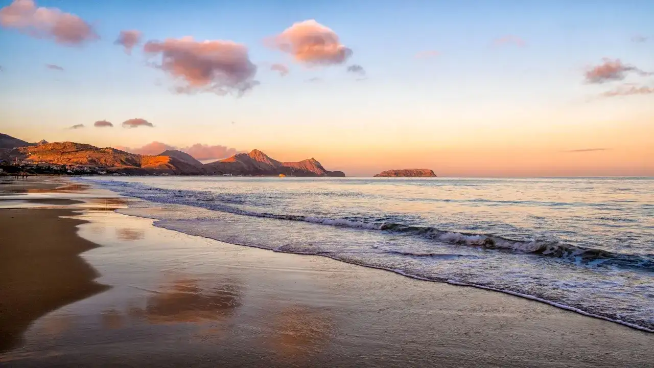 Sonnenuntergang am Strand mit sanften Wellen. Ein idyllischer badeurlaub im winter, mit Bergen im Hintergrund und rosa Wolken am Himmel.