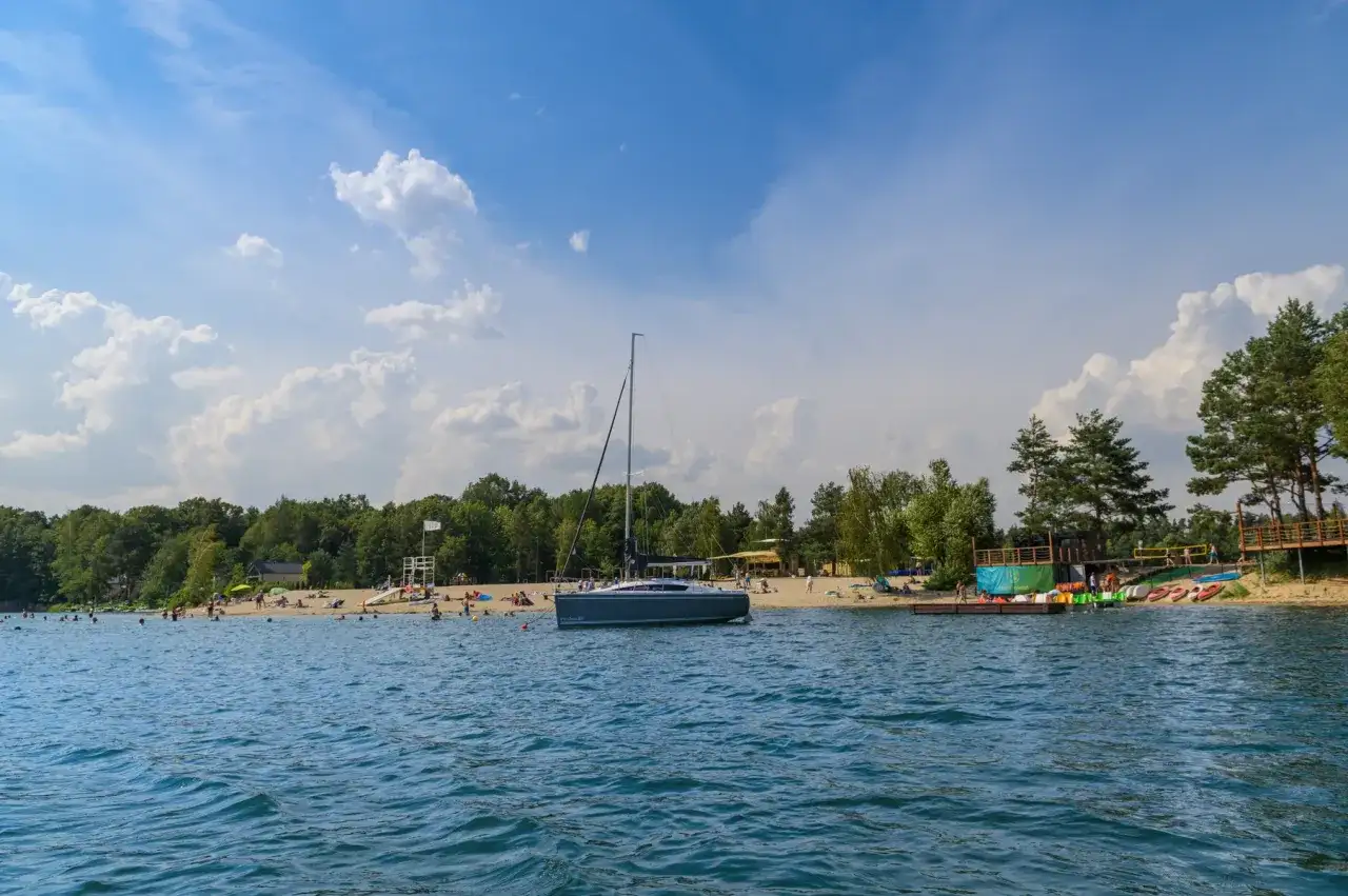 Żaglówka cumuje przy piaszczystej plaży nad jeziorem Głębocko Opolskie. Ludzie odpoczywają i kąpią się w słoneczny dzień.