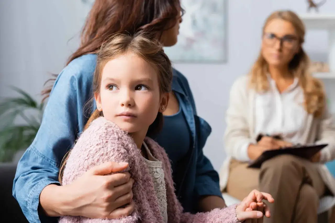 Dziewczynka w ramionach matki, zaniepokojona, szuka wsparcia. W tle terapeuta. Obrazuje skutki niepraktykowania wiary przez rodziców dla duchowości dziecka.