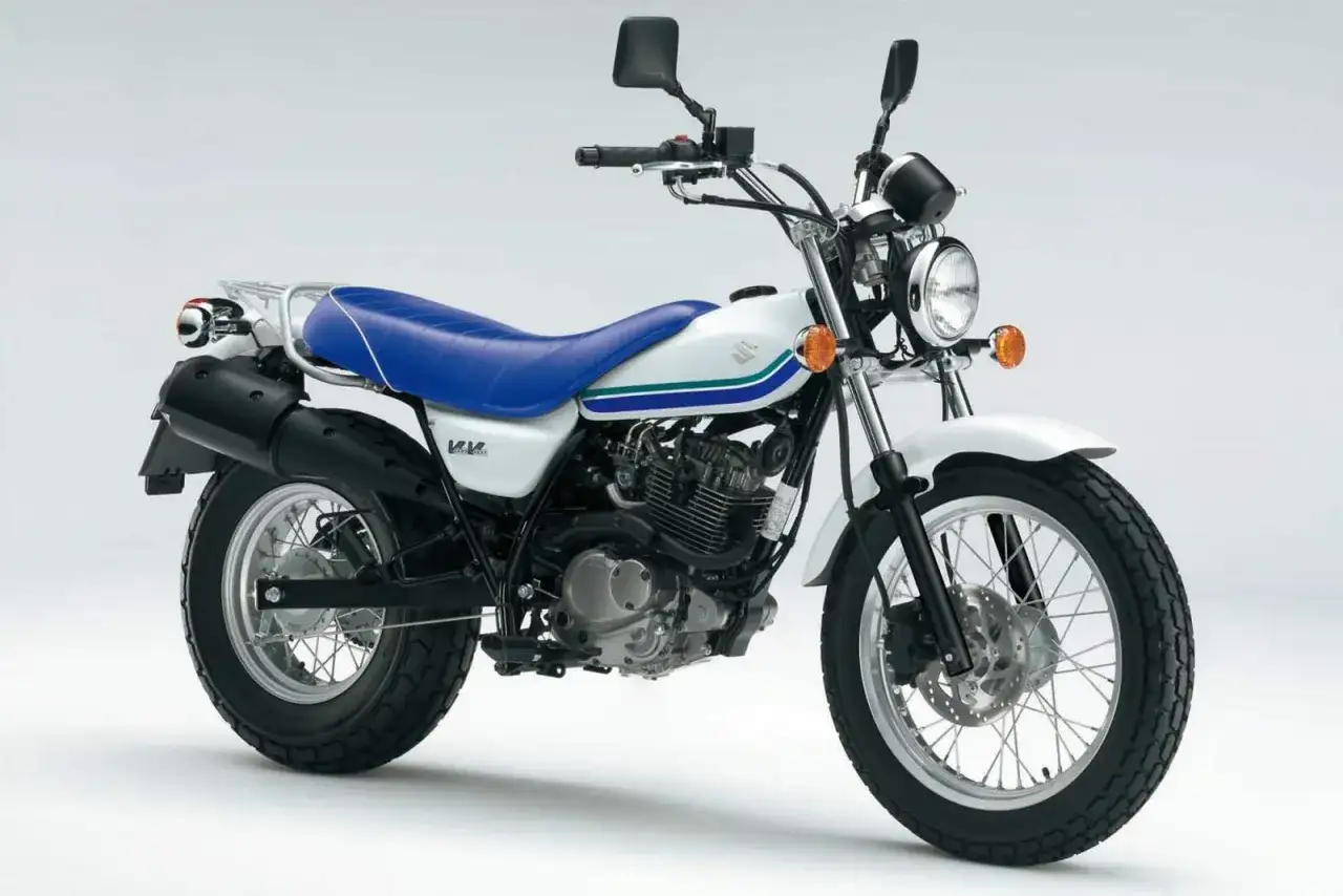 Biały motocykl Suzuki Van Van z niebieskim siedzeniem i zielono-niebieskimi pasami na baku. Idealny na miejskie wyprawy.