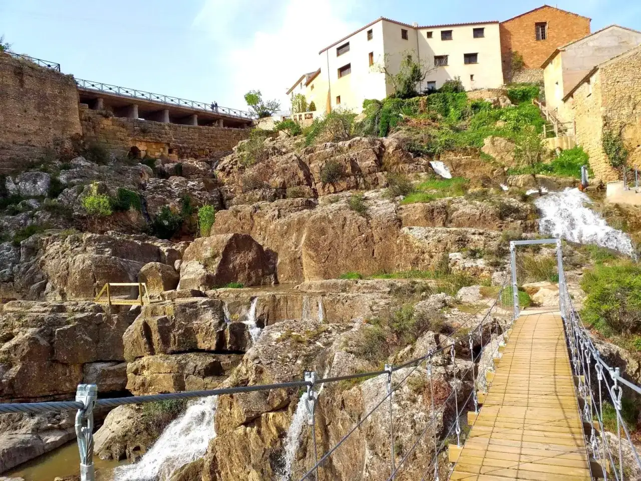Puente colgante sobre cascadas en Almonacid de la Cuba, con casas tradicionales en la ladera rocosa.