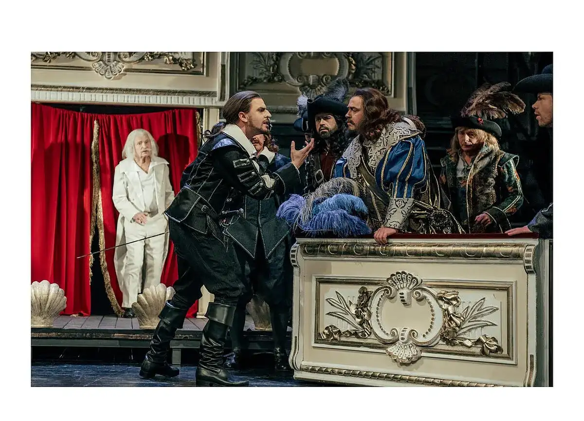 Aktorzy w strojach z epoki na scenie, w tym mężczyzna w czarnym stroju, przypominający Cyrano de Bergerac, gestykuluje do towarzyszy.
