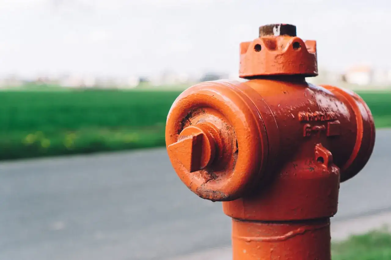 Pomarańczowy hydrant stoi blisko drogi, a za nim widać zielone pole. Odległość hydrantu od ogrodzenia jest niewielka.