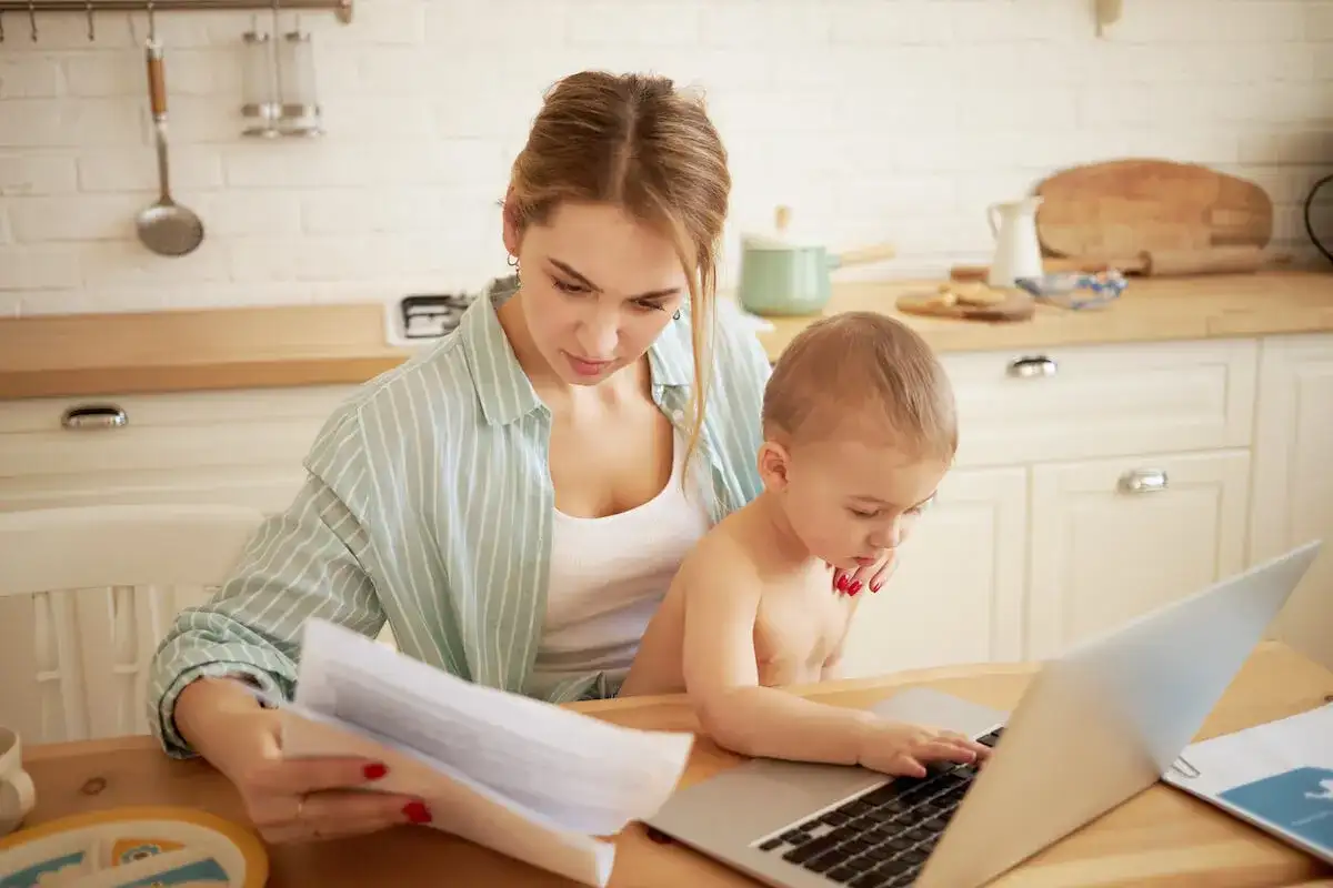Mama pracuje przy laptopie, a dziecko próbuje jej pomóc. To codzienność, gdy macierzyński ile trwa.