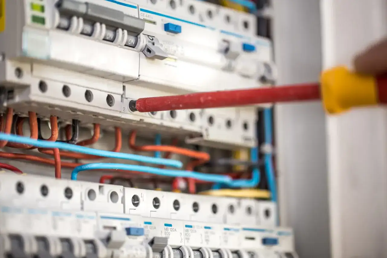 Ręka z czerwonym śrubokrętem pracuje przy panelu z wyłącznikami i kolorowymi przewodami. To instalacja niskoprądowa.